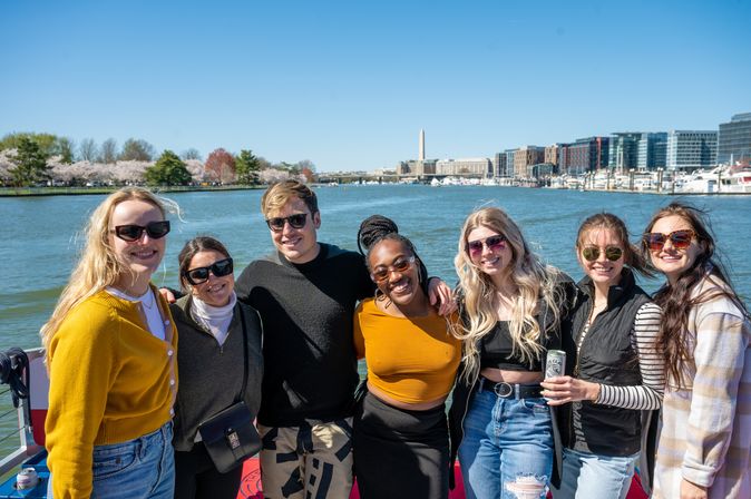 Group of seven smiling young adults on a sunny boat cruise in Washington, D.C., with cherry blossoms, a marina and the Washington Monument visible across the water.