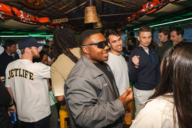 Crowded tiki-style boat bar at night — friends chatting and laughing with cocktails under a thatched roof and green neon lighting.
