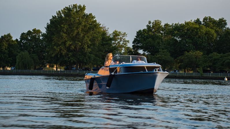 Small blue motorboat with two people cruising on a calm lake at golden hour, tree-lined shoreline and gentle rippling water.