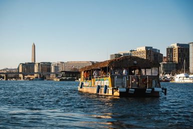 Tiki-style party boat cruising the Potomac River with the Washington Monument and Washington, D.C. waterfront skyline in warm golden-hour light.