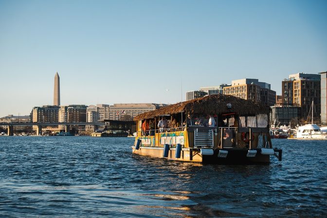 Tiki-style party boat cruising the Potomac River with the Washington Monument and Washington, D.C. waterfront skyline in warm golden-hour light.