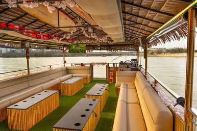 Tiki-style riverboat interior with long beige cushioned benches, bamboo tables with cup holders on green turf, hanging floral garlands and a thatched roof, steering helm and waterfront river view