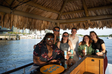 Six friends smiling under a thatched tiki roof at a boat-side bar in a sunny marina, holding canned drinks with yachts and city buildings in the background.