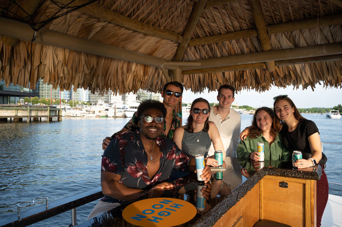 Six friends smiling under a thatched tiki roof at a boat-side bar in a sunny marina, holding canned drinks with yachts and city buildings in the background.