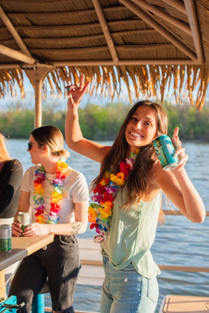 Smiling woman wearing colorful leis holding a canned drink under a tiki-style canopy on a sunny pontoon boat by the lake