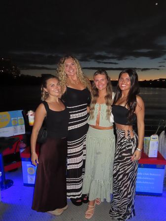 Four smiling friends in stylish summer outfits posing on a waterfront deck at dusk with city lights and a moody sunset sky in the background.