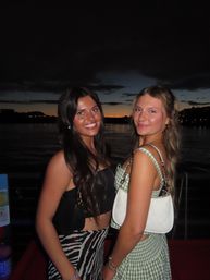 Two smiling women in stylish summer outfits posing on a waterfront promenade at sunset with a calm river and city lights in the background
