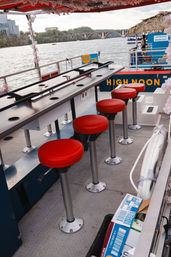 Riverside party boat bar with a row of red swivel stools and a shuffleboard-style counter overlooking a stone-arch bridge and river in Washington, D.C.