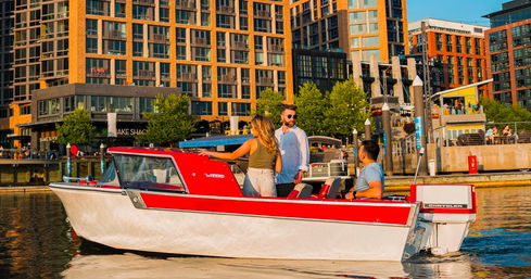 Three people enjoy a red-and-white vintage motorboat on a sunny city marina, cruising past modern waterfront apartments and dockside restaurants.