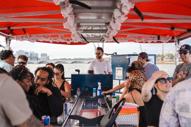 Festive group on a red-canopied riverboat cruise with floral garlands, outdoor bar and bartender/DJ, enjoying drinks and music as a city skyline and bridge pass by