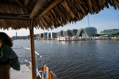 View from a thatched-roof boat across a Washington, D.C. waterfront marina with docked yachts, modern glass buildings, and the Washington Monument on the skyline.