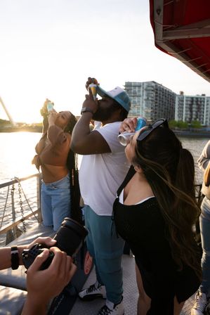 Three people on a boat deck chugging canned drinks at sunset with an urban waterfront and apartment skyline in the background.