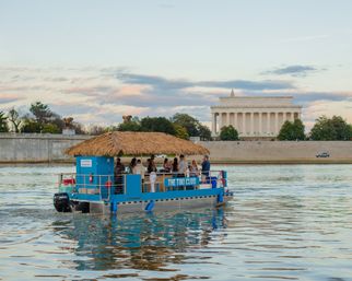 Tiki-style party pontoon with a straw roof cruising the Potomac at sunset, water reflections and the Lincoln Memorial on the National Mall in Washington, D.C.