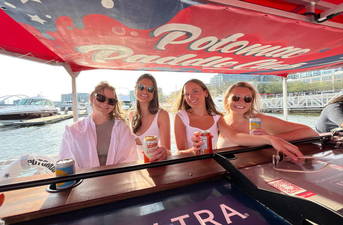 Four friends smiling on a pontoon boat under a red canopy, holding canned drinks at a Potomac River waterfront marina with yachts and city buildings in the background.