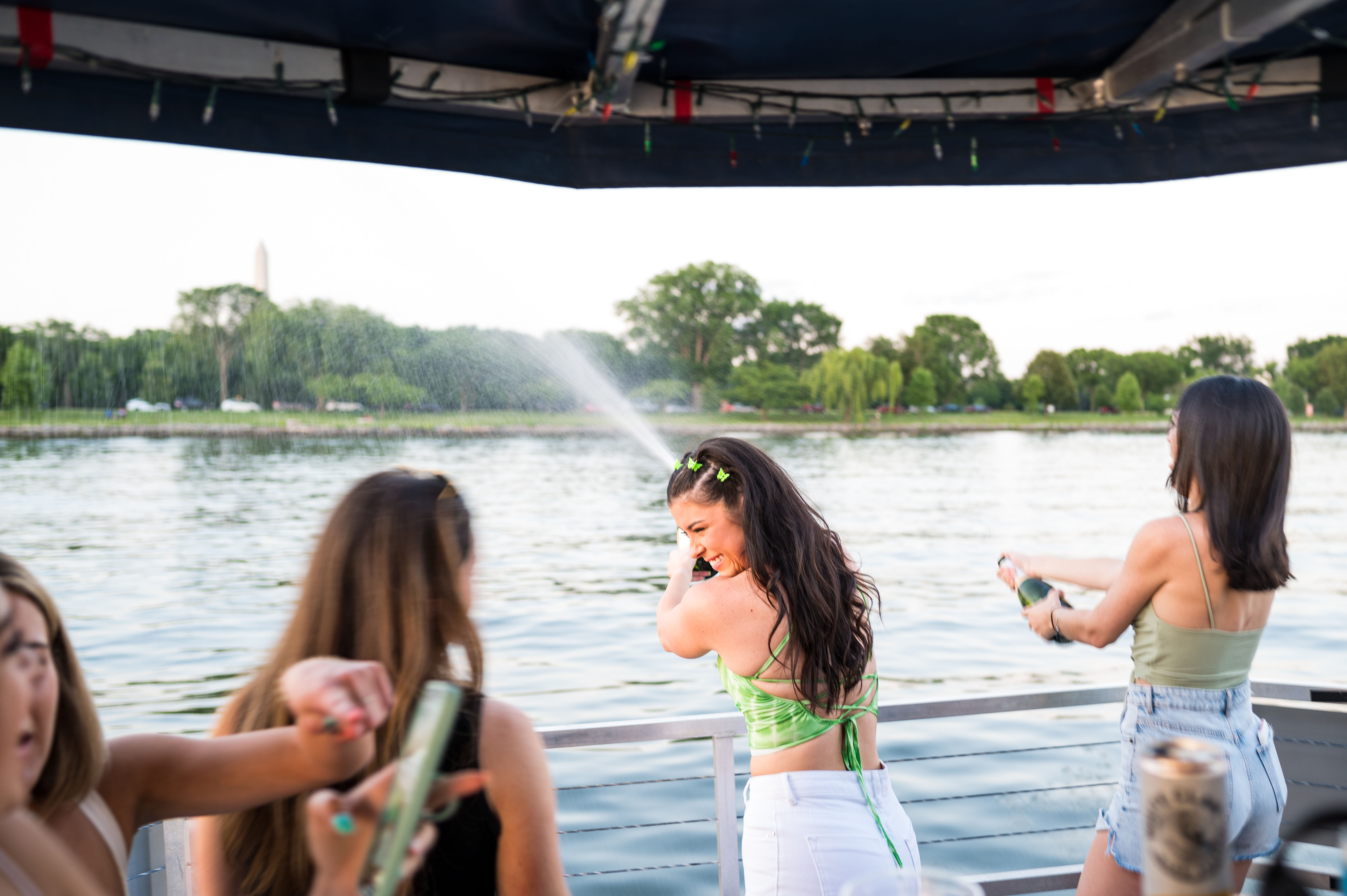 Friends on a boat near the Washington Monument in Washington, D.C., laughing as champagne sprays across the water on a sunny summer evening.
