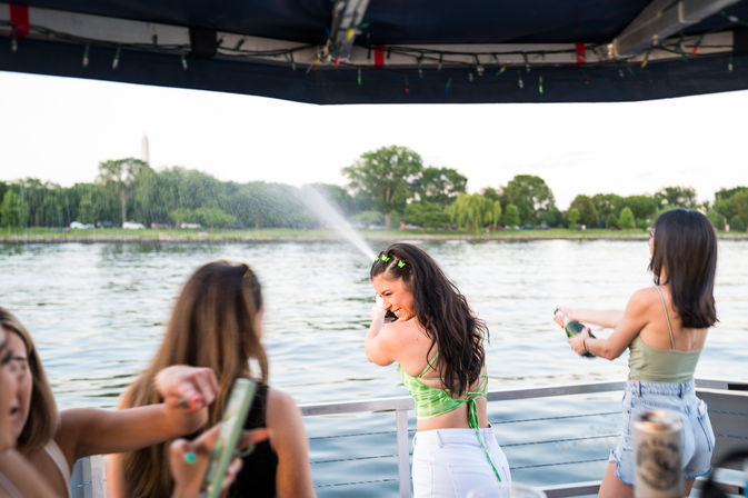 Friends on a boat near the Washington Monument in Washington, D.C., laughing as champagne sprays across the water on a sunny summer evening.