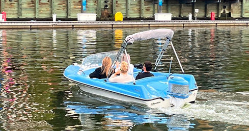 Blue motorboat with three passengers cruising near a wooden dock, leaving ripples and colorful reflections on calm harbor water.