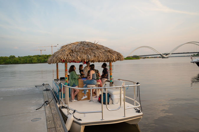 Group of friends on a tiki‑themed pontoon boat bar at a riverside marina at sunset, thatched roof over the bar, calm water and arched bridge in the background.