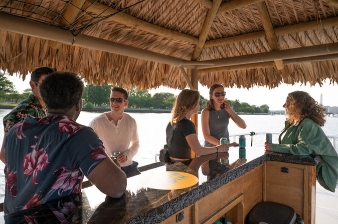Smiling friends share drinks at a tiki-style boat bar on the Washington, D.C. waterfront, with the Washington Monument and river in the background.