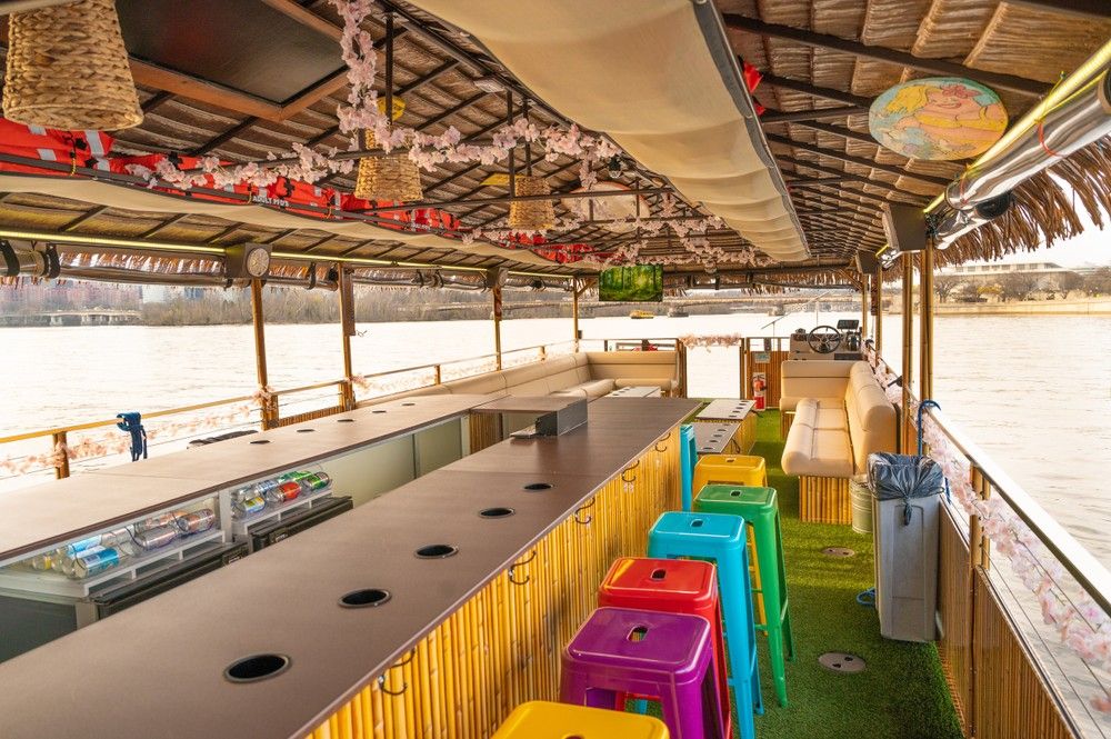 Tiki-style riverboat bar interior on a calm river with a bamboo counter and built-in coolers, rainbow plastic bar stools on artificial turf, hanging floral garlands and woven lamps, cushioned bench seating and a captain's helm with waterfront city skyline visible through open sides.