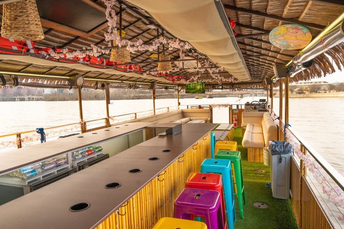 Tiki-style riverboat bar interior on a calm river with a bamboo counter and built-in coolers, rainbow plastic bar stools on artificial turf, hanging floral garlands and woven lamps, cushioned bench seating and a captain's helm with waterfront city skyline visible through open sides.