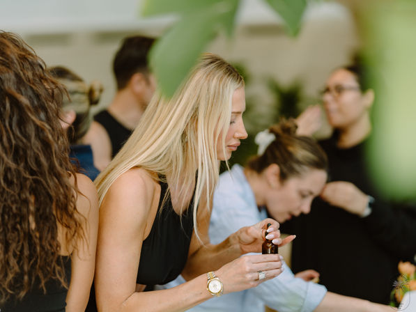 Blonde woman in a black sleeveless top examines a small amber dropper bottle while other women sample skincare or essential oils at an indoor wellness event.