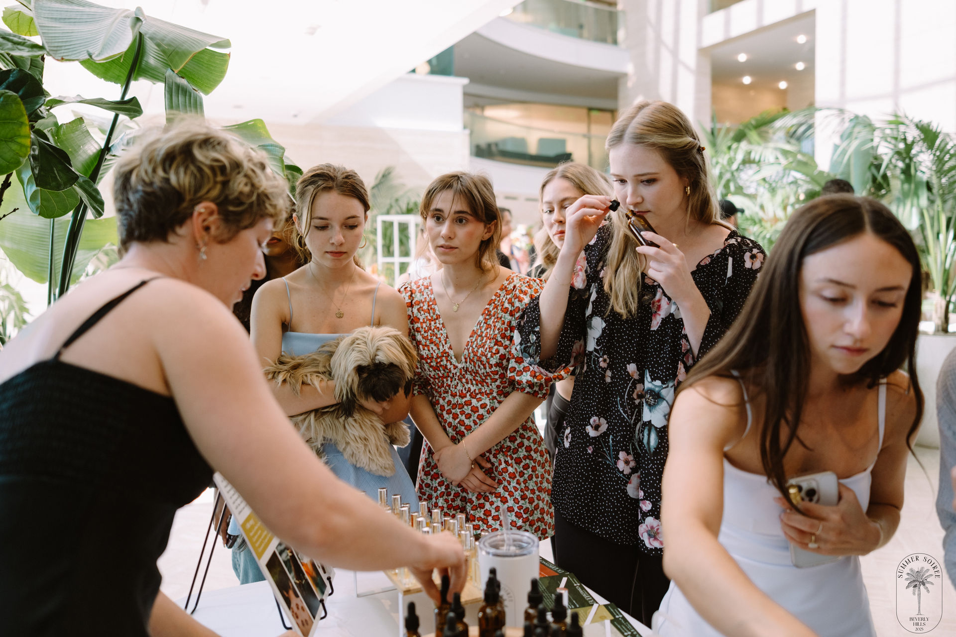 Young women at a bright, plant-filled indoor atrium pop-up shop sampling perfume oils — one sniffs a tester, another holds a small fluffy dog while a vendor arranges glass bottles.