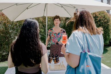 Outdoor market booth under a white umbrella with a vendor in a colorful blouse talking to two shoppers; a table displays amber dropper bottles and sample vials while one shopper holds a pink cocktail in a wine glass.