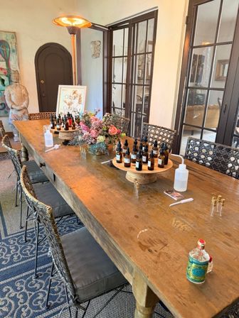 Sunlit rustic wooden table set for a perfume/essential oil blending workshop with rows of amber dropper bottles on rotating wooden displays, a floral centerpiece, hand sanitizer pump and woven chairs in an airy room with French glass doors.