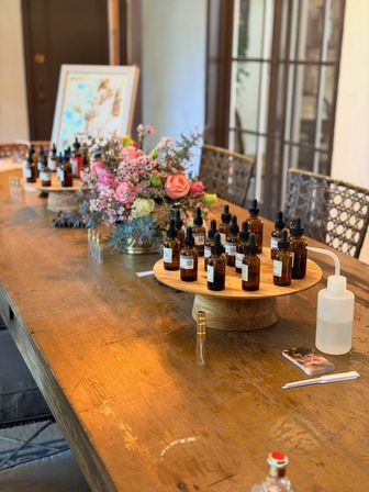 Sunlit studio table set for an aroma workshop: amber dropper bottles on wooden risers, perfume sample vials, squeeze bottle and a pink floral centerpiece.