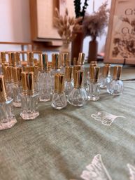 Rows of small clear glass perfume bottles with shiny gold caps arranged on a green cloth tabletop, with blurred dried flower vases and soft indoor decor in the background — elegant boutique-style display