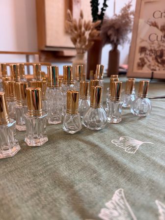 Rows of small clear glass perfume bottles with shiny gold caps arranged on a green cloth tabletop, with blurred dried flower vases and soft indoor decor in the background — elegant boutique-style display
