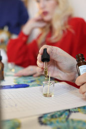 Close-up of hands dropping golden essential oil from a glass dropper into a small beaker with a toothpick, notebook and pen on the table during an aromatherapy DIY workshop.