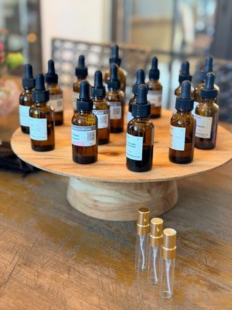 Amber glass dropper bottles with labels arranged on a round wooden display stand and three clear perfume sample vials with gold caps on a rustic tabletop — apothecary-style fragrance sampling display in a boutique.