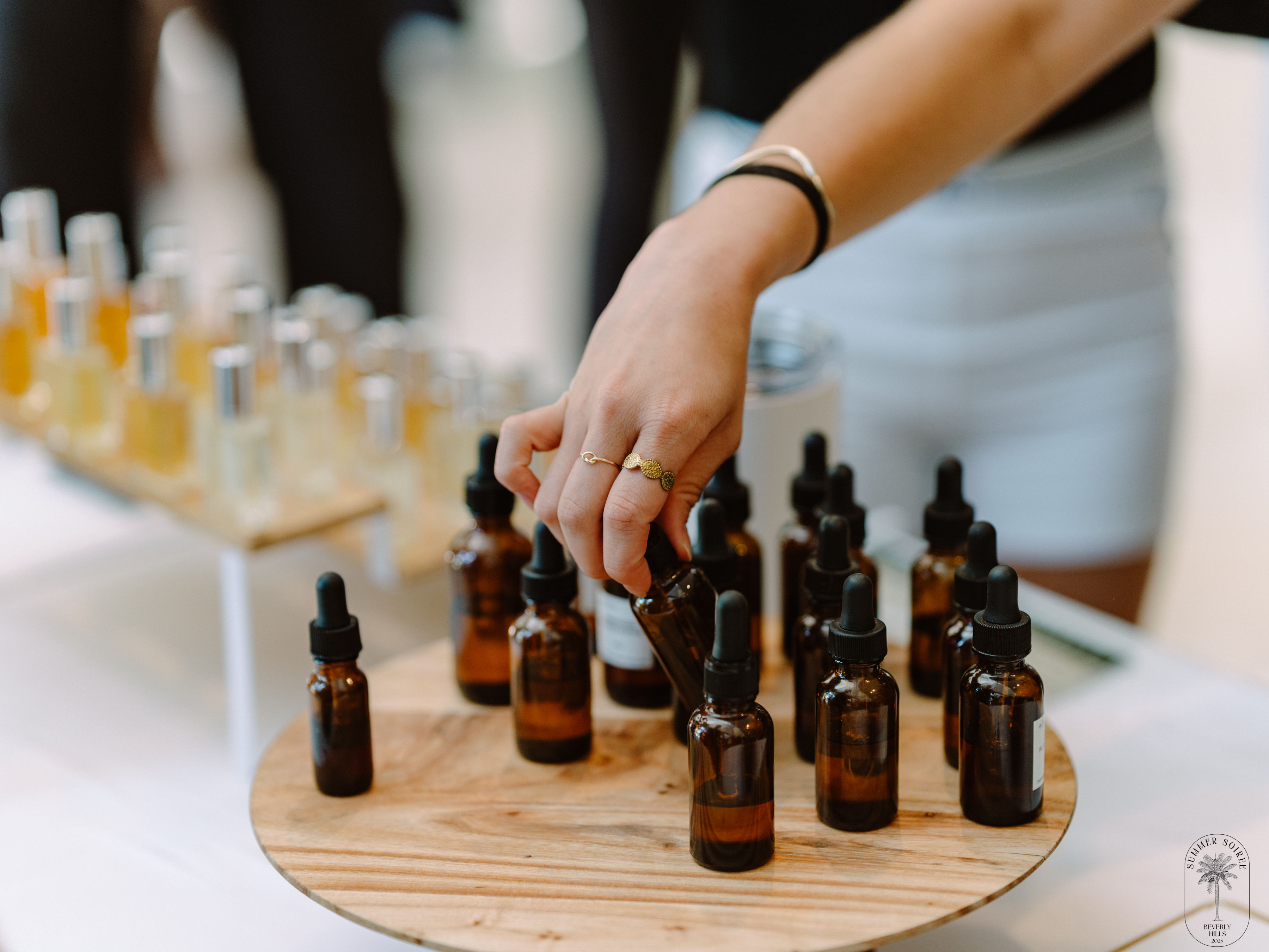 Hand reaching to pick an amber glass dropper bottle from a wooden display of essential oil and serum bottles at a market stall