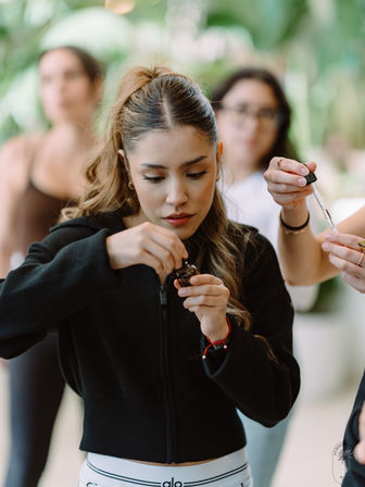 Focused woman in activewear opens an amber essential-oil bottle while another hand uses a dropper during an indoor aromatherapy wellness workshop.