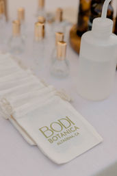 Natural cotton drawstring pouches with green logo beside small glass perfume vials with gold caps and a frosted squeeze bottle on a white tablecloth.