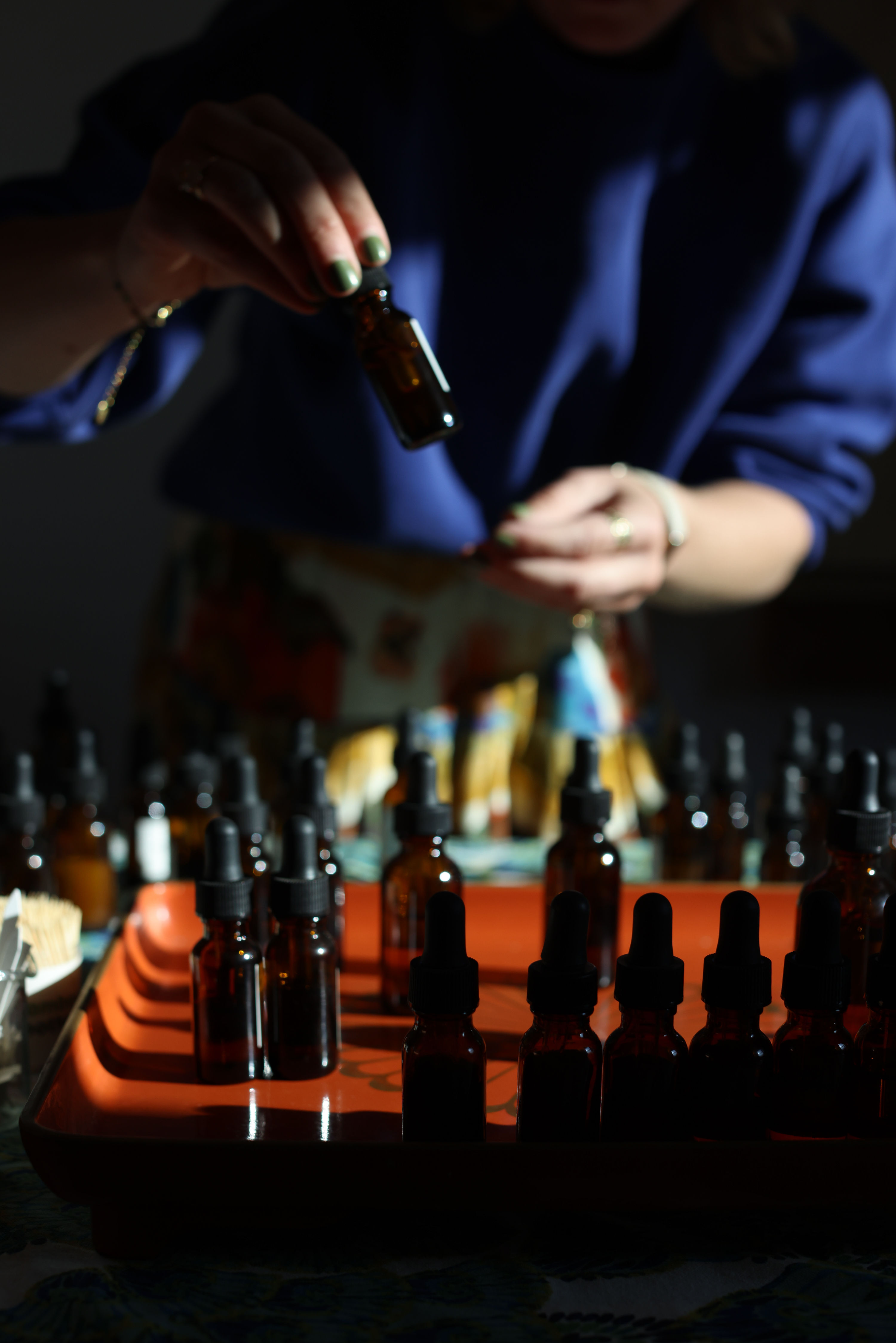 Hands lifting an amber glass dropper bottle from an orange tray of amber droppers on a studio table, aromatherapy essential oils in moody light.