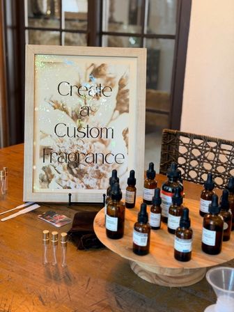 Inviting indoor fragrance bar with a wooden-framed sign reading 'Create a Custom Fragrance', amber dropper bottles, small glass sample vials and a wooden display tray on a rustic table.