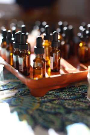 Backlit amber glass dropper bottles with black caps arranged on a wooden tray atop a colorful patterned tablecloth — warm aromatherapy/apothecary display.