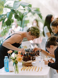 Customers sampling essential oils at a sunlit, plant-filled pop-up market stall — amber dropper bottles on a wooden display with fresh flowers and testers.