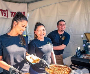 Three smiling servers in black shirts scoop shredded meat into tacos from a steaming pan at an outdoor food festival booth