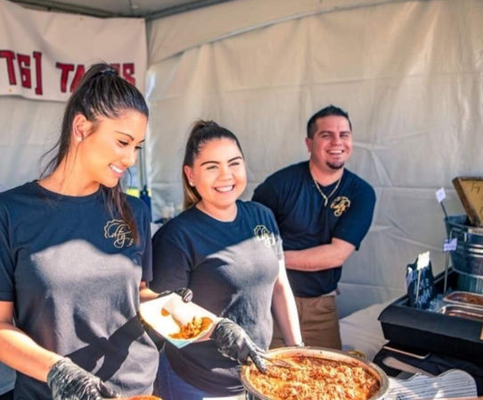 Three smiling servers in black shirts scoop shredded meat into tacos from a steaming pan at an outdoor food festival booth