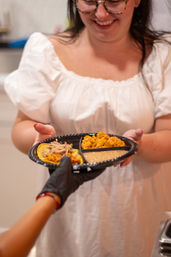 Smiling person in a white dress accepting a black compartment plate with shredded meat tacos, seasoned yellow rice and beans from a gloved server