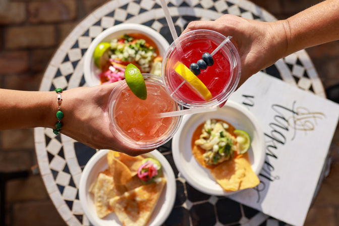 Overhead shot of two colorful iced cocktails (one pink with a lime wedge, one red with lemon and blueberries) clinking above plates of tacos and chips on a mosaic outdoor patio table