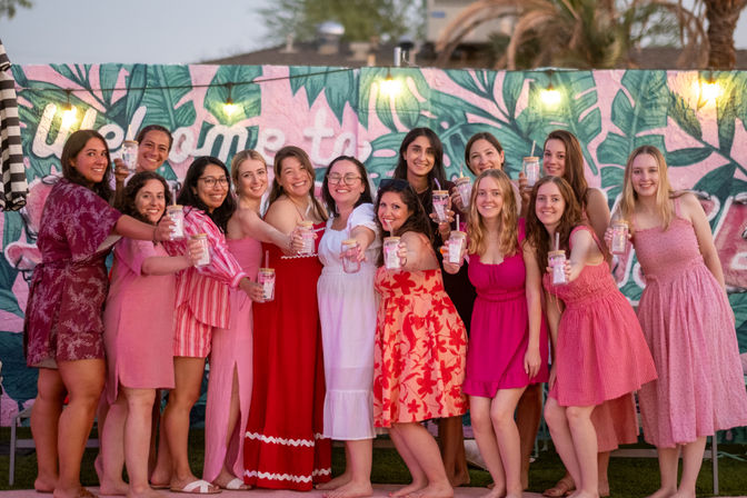 Group of friends in pink and red dresses cheering with mason-jar drinks in front of a colorful tropical palm-leaf mural and string lights on an outdoor patio