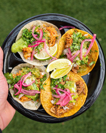 Hand holding a black plate of four vibrant street tacos—corn and flour tortillas with shredded meat, green salsa, chopped onions, cilantro, pickled red onions and lime wedges on a grass background