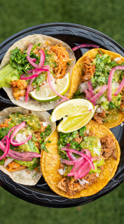 Hand holding a black plate of four vibrant street tacos—corn and flour tortillas with shredded meat, green salsa, chopped onions, cilantro, pickled red onions and lime wedges on a grass background