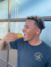 Smiling young man outdoors enjoying a crunchy taco with greens, standing by a restaurant window on a sunny day in a casual street-taco scene.