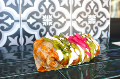 Close-up of a grilled Mexican-style burrito drizzled with salsa verde and crema, topped with pickled red onions and cilantro, placed on glossy blue tiles with a patterned tile backsplash.